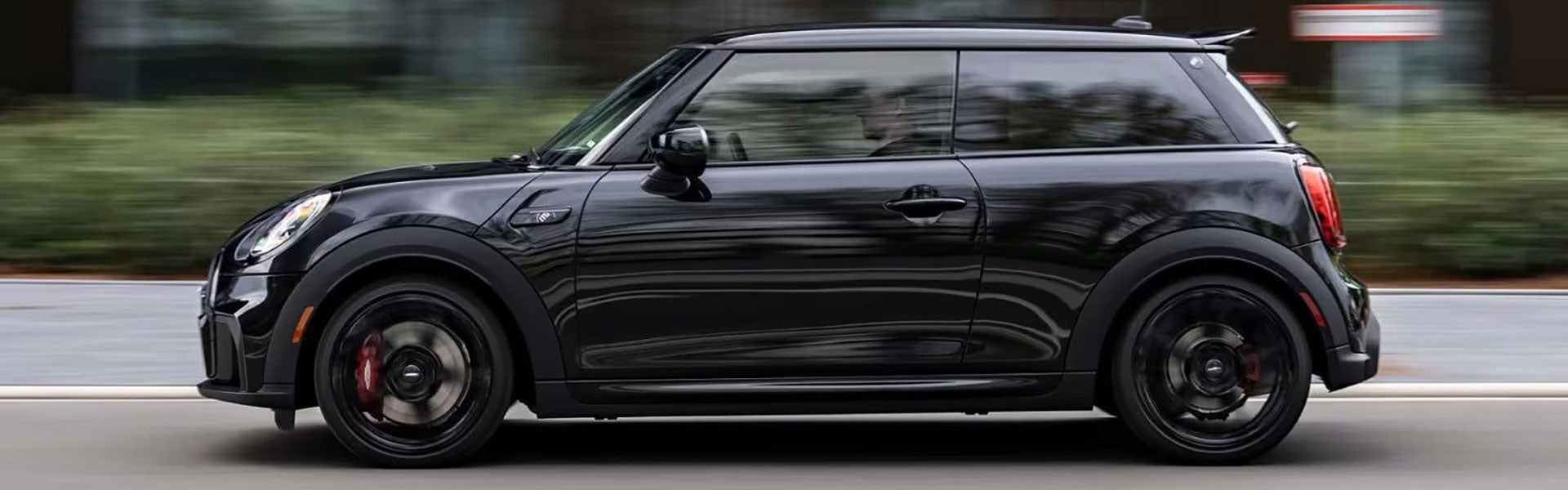 Side view of a MINI John Cooper Works 1to6 Edition vehicle driving alone on a street facing left, with bushes in the background along and the vehicle's surroundings blurred out.