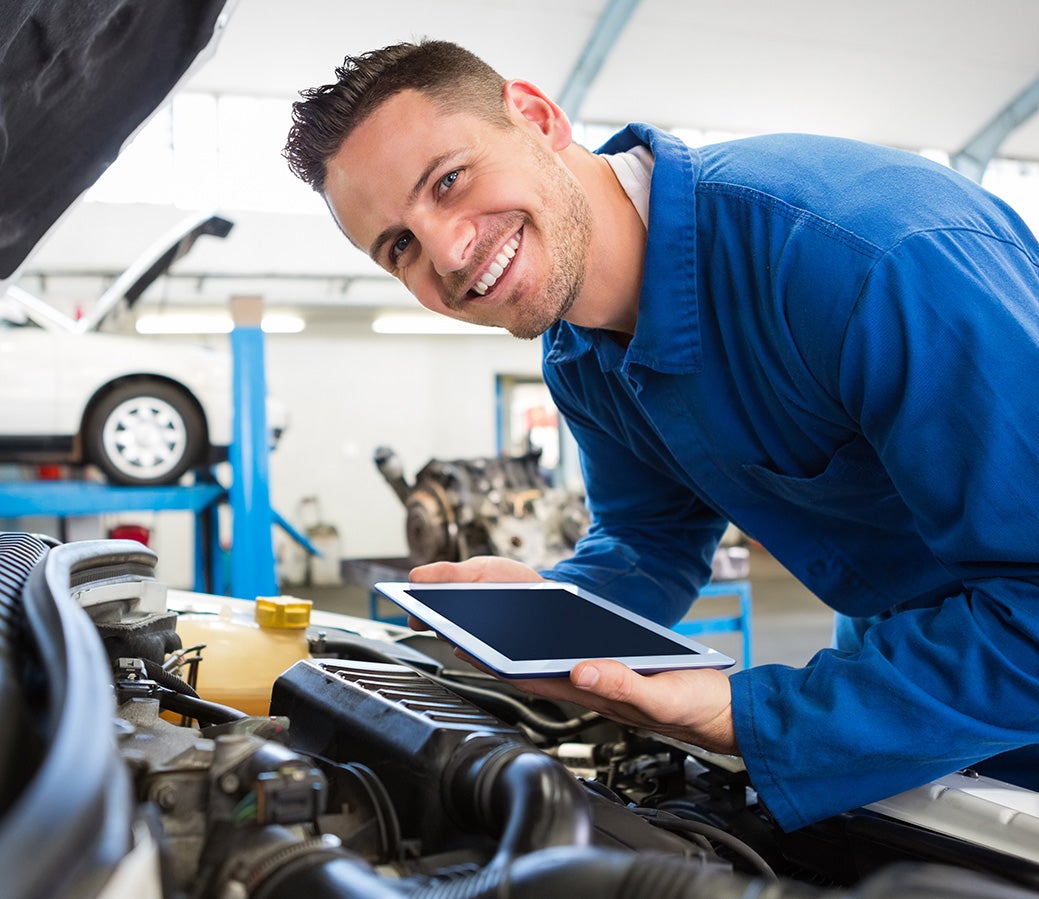 Smiling technician servicing a car engine while holding a tablet.